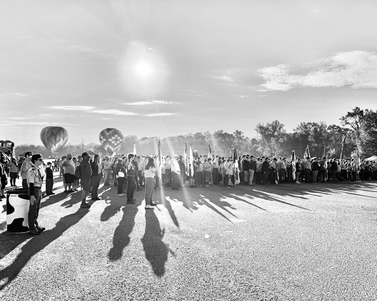 A black and white photo of a crowd of people standing in a field with hot air balloons in the background.