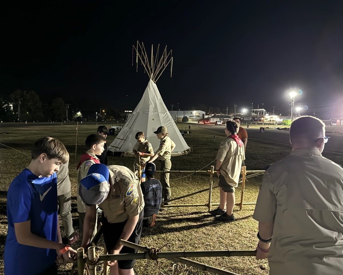 A group of boy scouts are standing in front of a teepee at night.