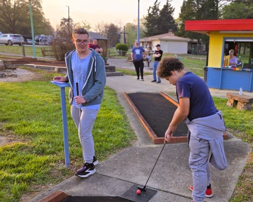 Two people playing mini golf outdoors, one lining up a putt near a colorful course booth.