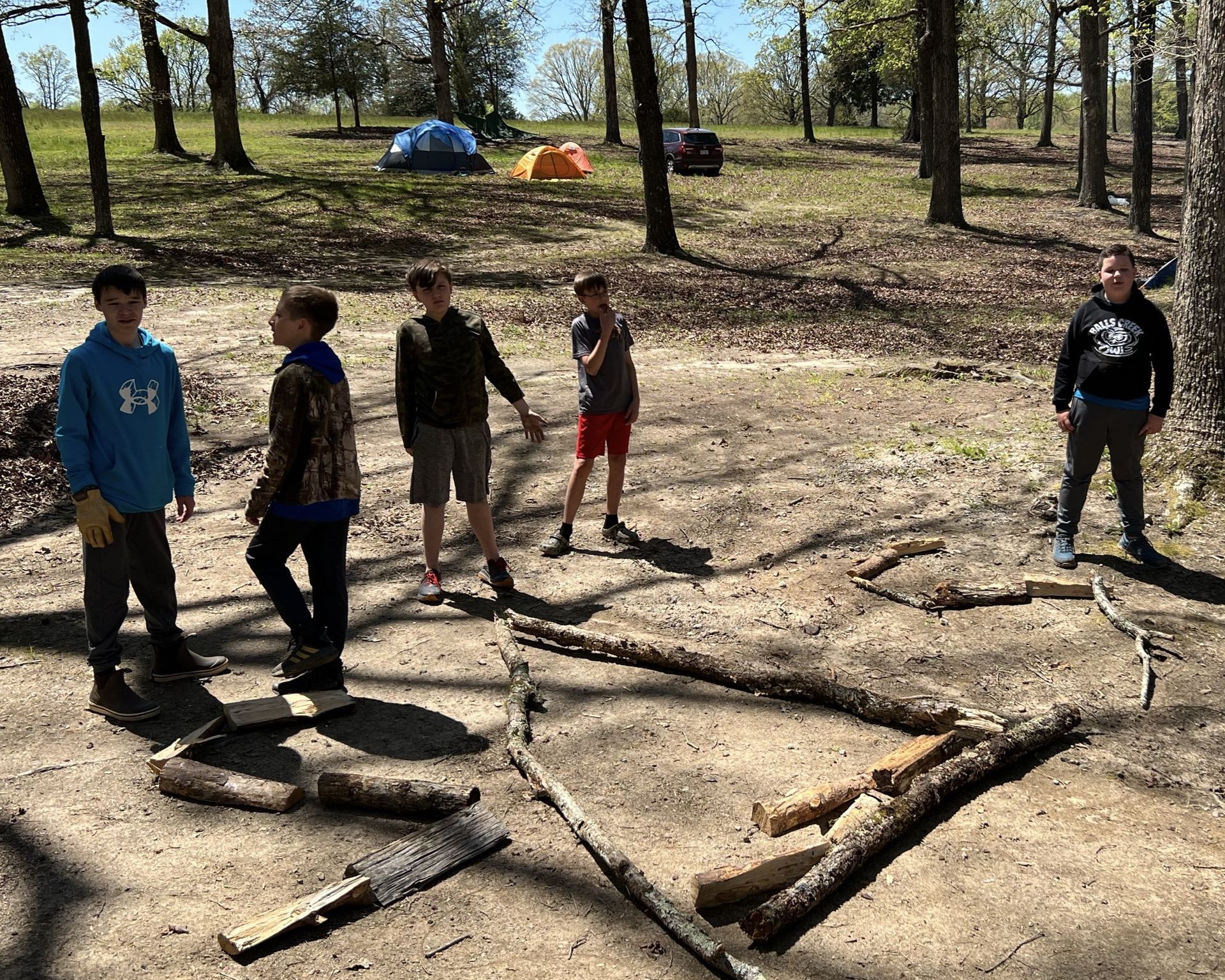 A group of young boys are standing around a fire pit in the woods.