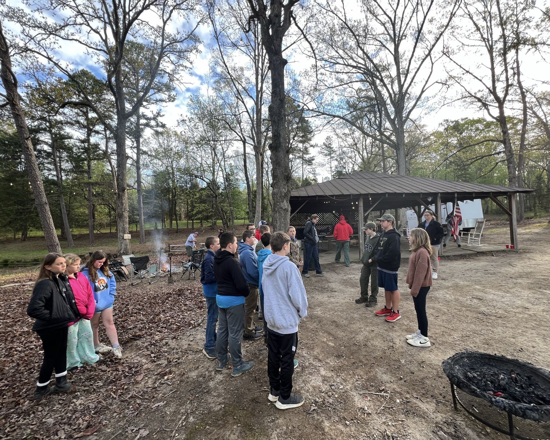 A group of scouts are standing around a fire pit in the woods.