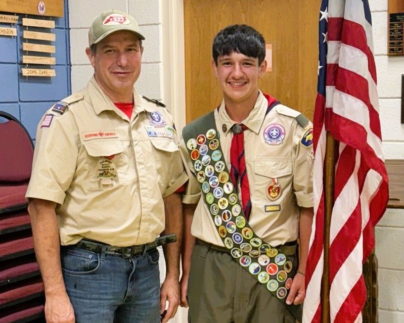 Two scouts in tan uniforms, one with merit badges, standing beside an American flag indoors.