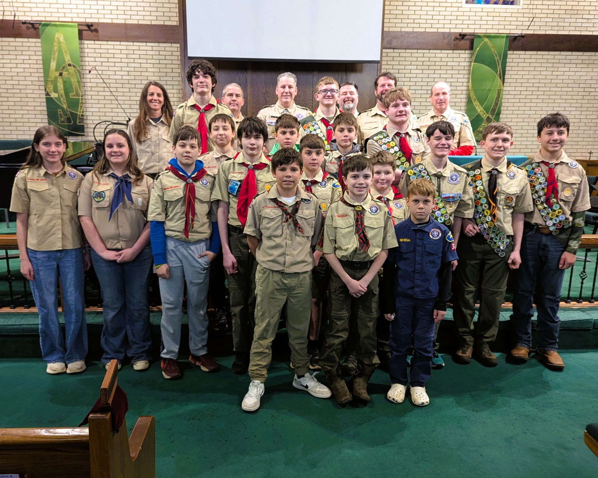 Scout troop group posing indoors in uniform at a ceremony, with a projection screen behind them.