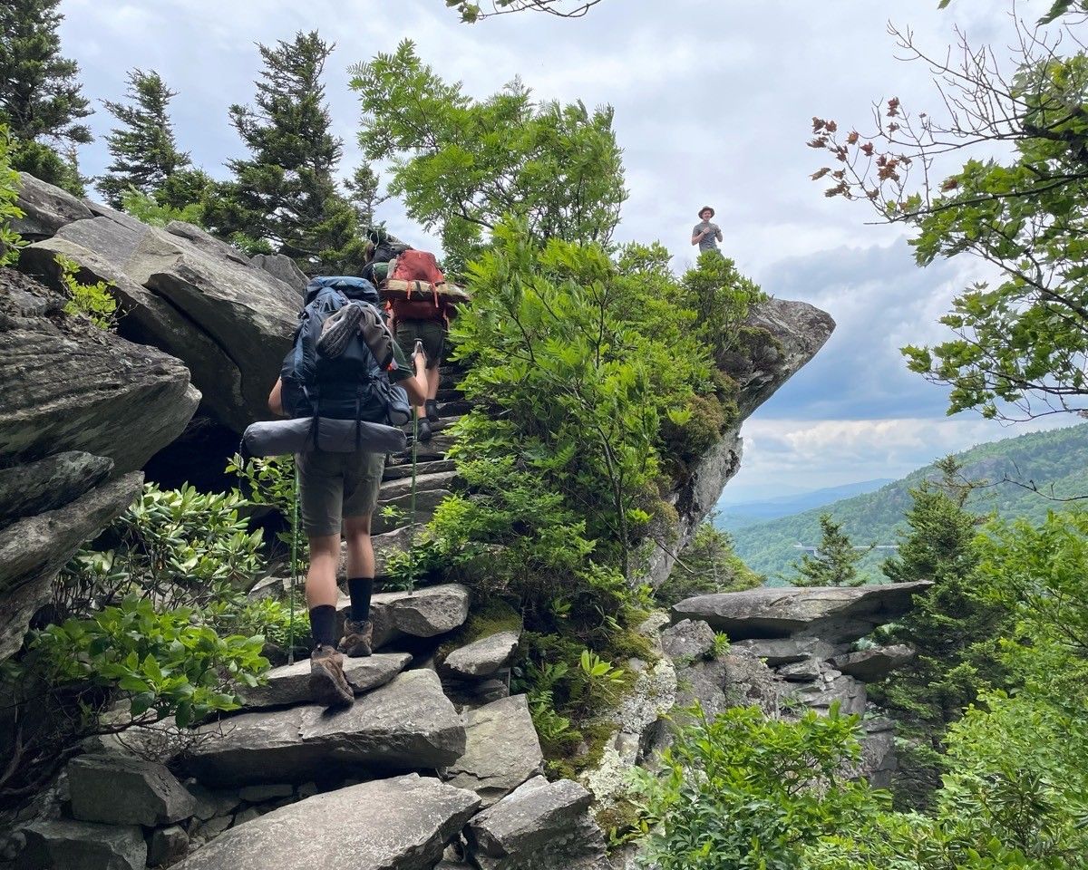 Hikers ascending rocky steps on a cliffside trail with green trees and mountain views