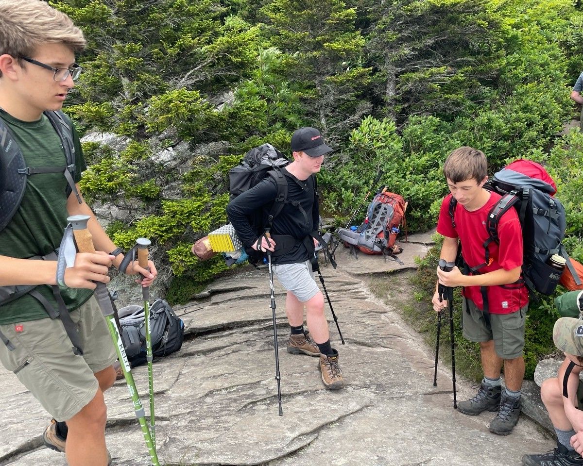 Three hikers with trekking poles climbing a rocky trail in a forested area