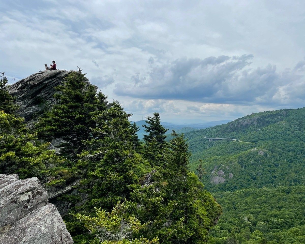 Rocky mountain overlook with two people on a peak above green forest and cloudy sky