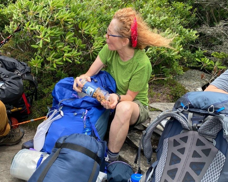 Person in green shirt tending to someone resting on hiking gear beside backpacks and greenery.
