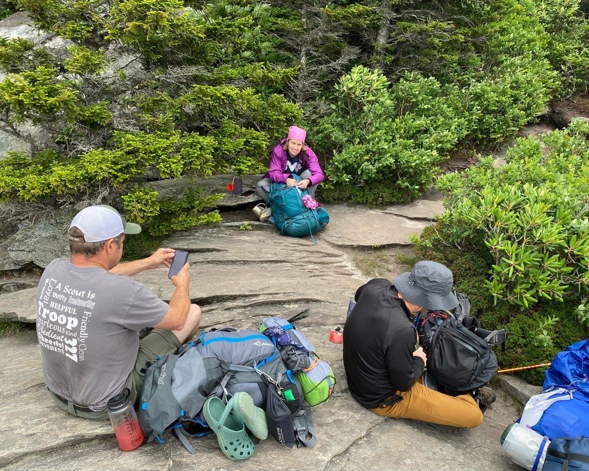 Hikers resting on a rocky trail beside green shrubs, with backpacks laid out and one person seated in pink and purple.