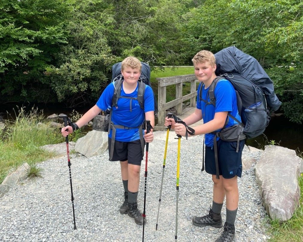 Two hikers in blue shirts with trekking poles stand on a gravel trail near large rocks and trees.