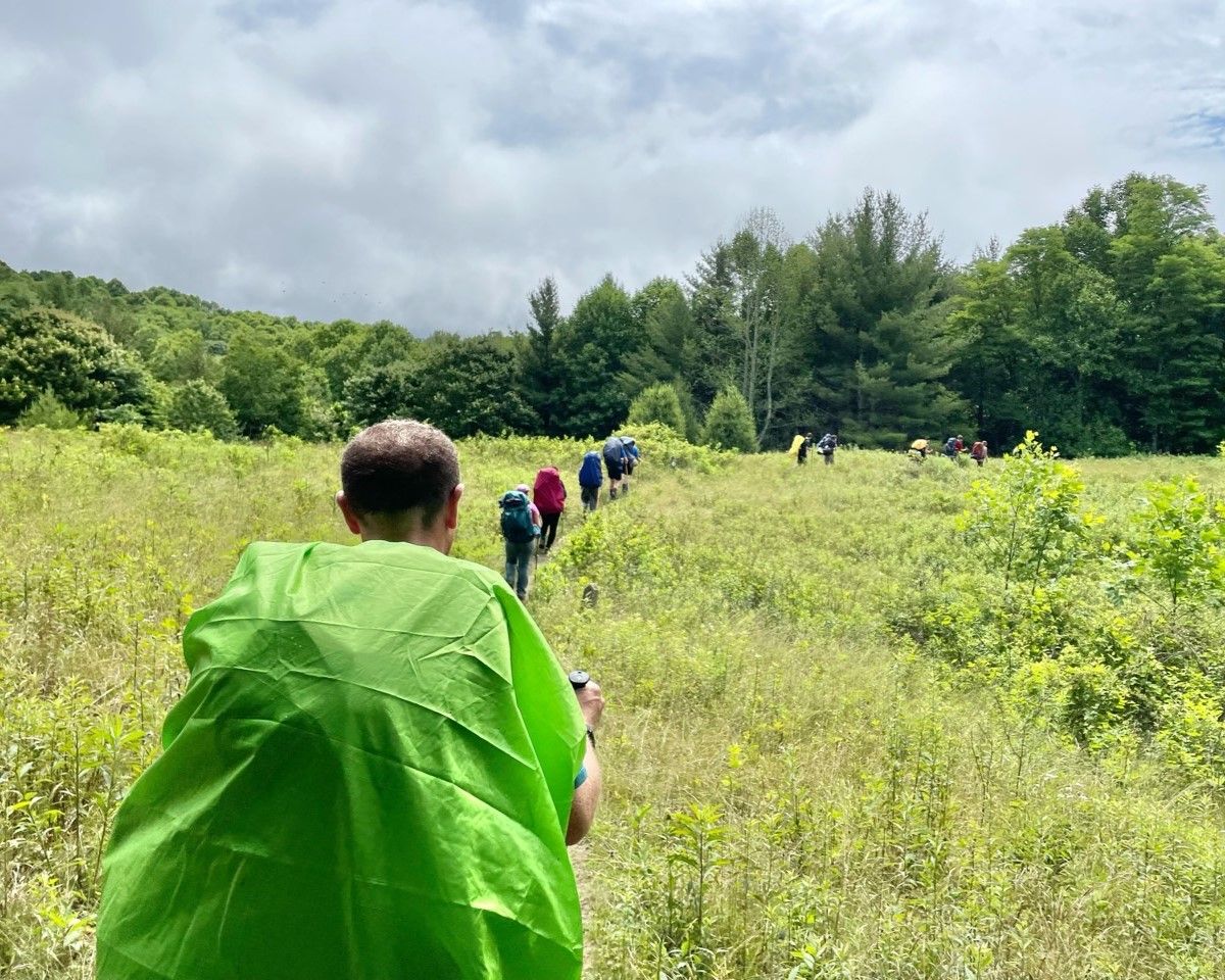 Hikers in a grassy field under cloudy skies, led by a person in a bright green poncho