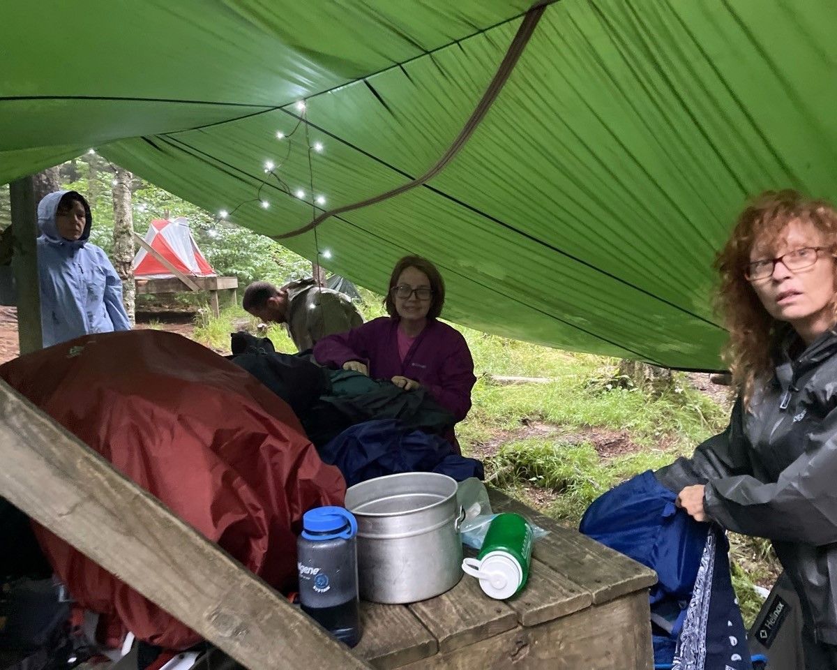 People sitting under a green tarp shelter with camping supplies and a large pot on a wooden table.