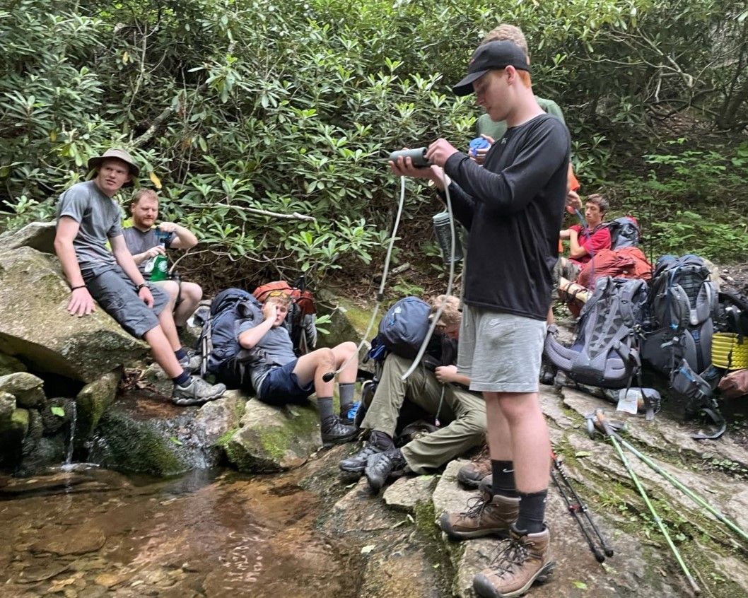 Hikers resting on rocks beside a shallow creek in a wooded area, with one person checking a phone.