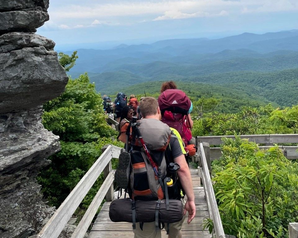 Hikers with backpacks walking on a wooden trail overlook above a lush mountain valley