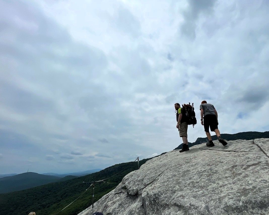 Two hikers stand on a rocky mountain summit under a cloudy sky.