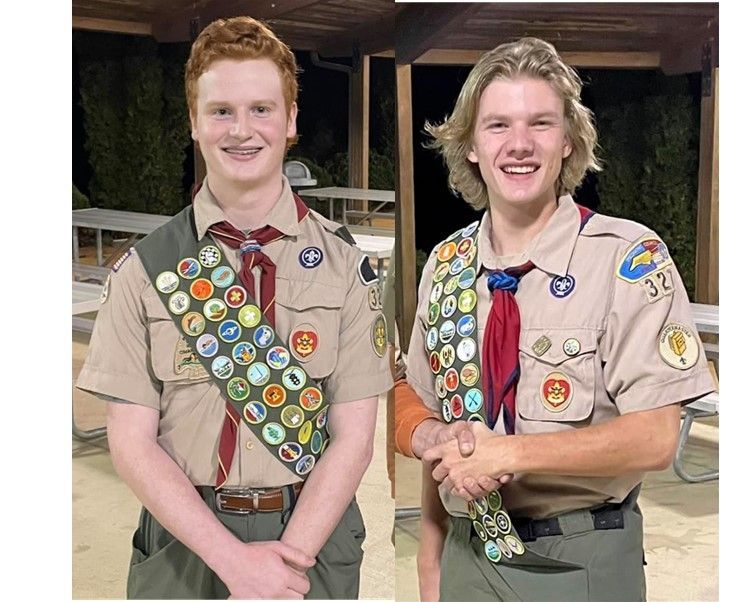 Two Scouts in tan uniforms with merit badges stand smiling indoors at night.