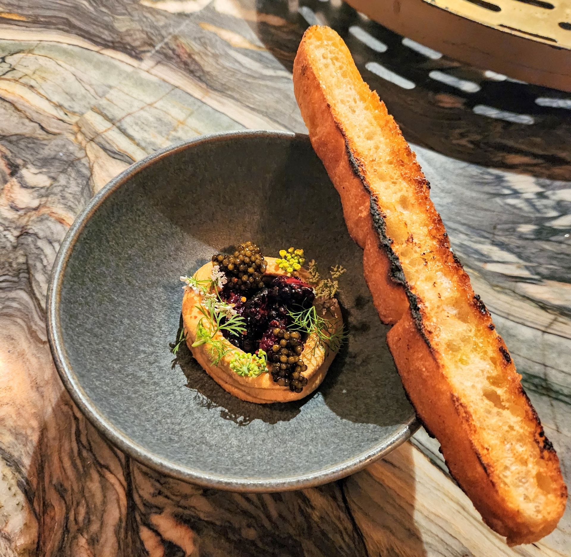 A bowl of food with toasted bread on a marble table. The dish has black caviar and herbs.