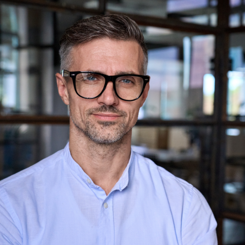 Man with glasses and a light blue shirt smiles in an office setting.