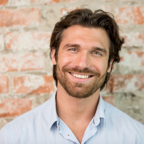 Man with brown hair and beard, smiling in front of a brick wall, wearing a light blue collared shirt.