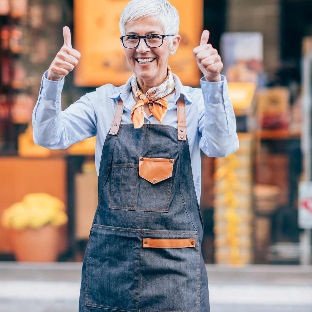 Woman in denim apron gives thumbs up outside a shop.
