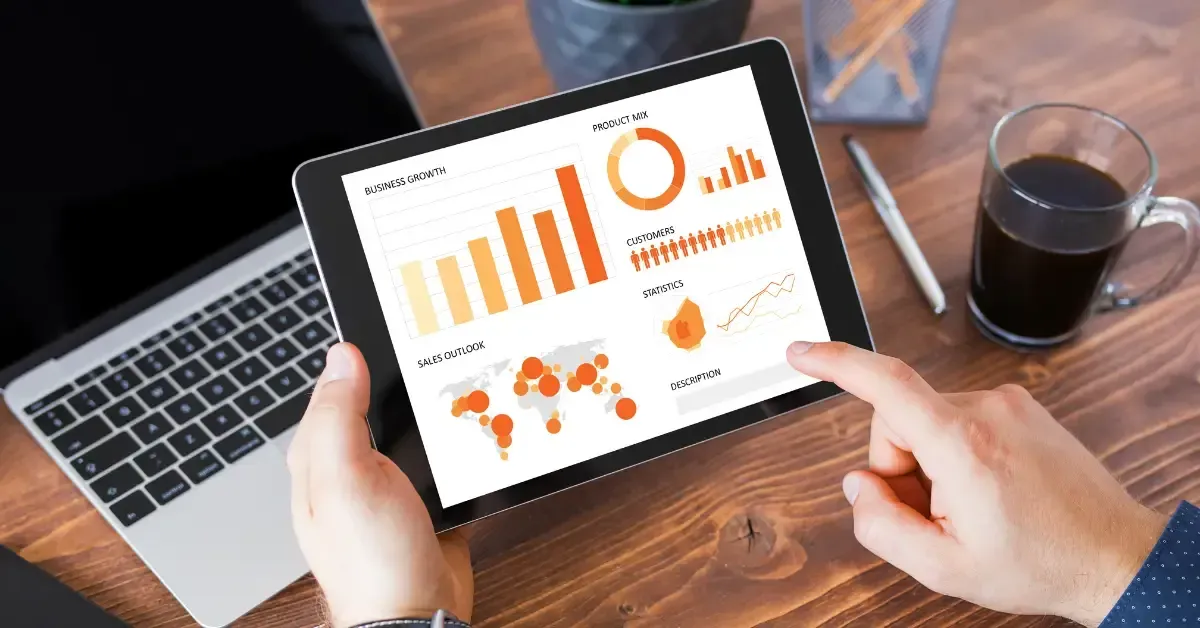 Hands holding a tablet displaying orange business charts on a desk with a laptop and coffee.