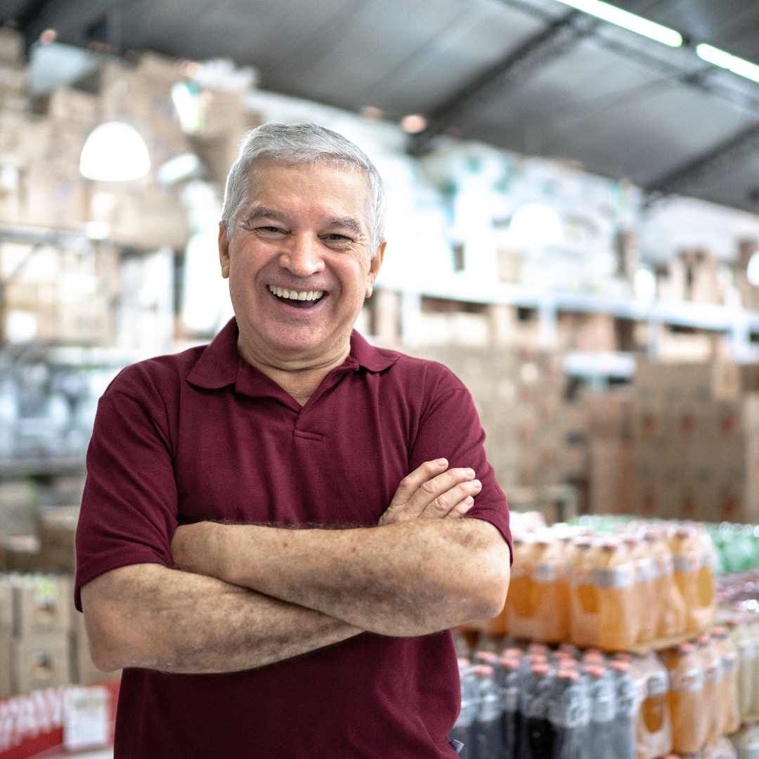 Man in maroon shirt smiles with arms crossed in a warehouse with drinks and products.
