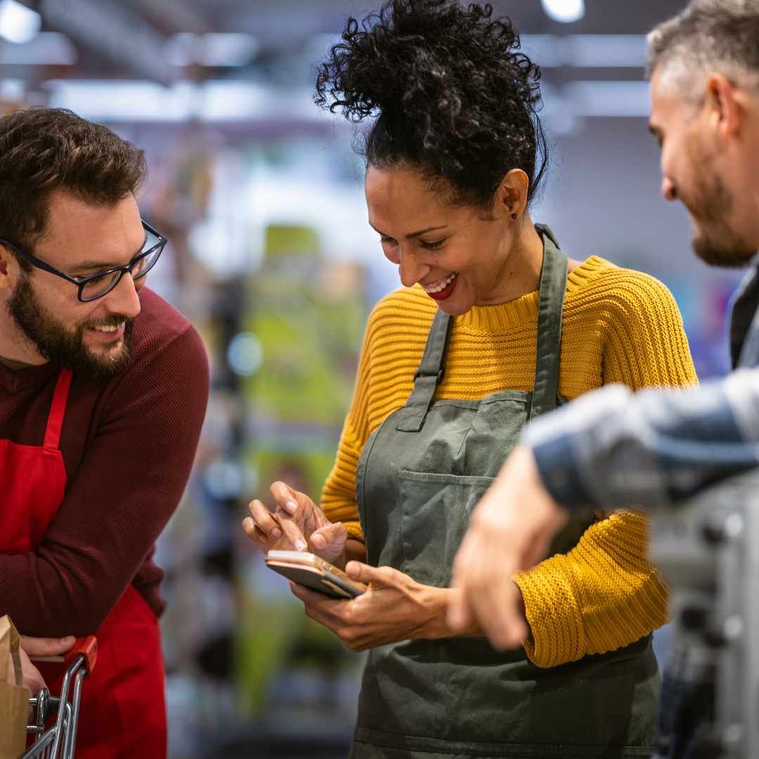 Three people in aprons smiling, looking at a phone in a grocery store.