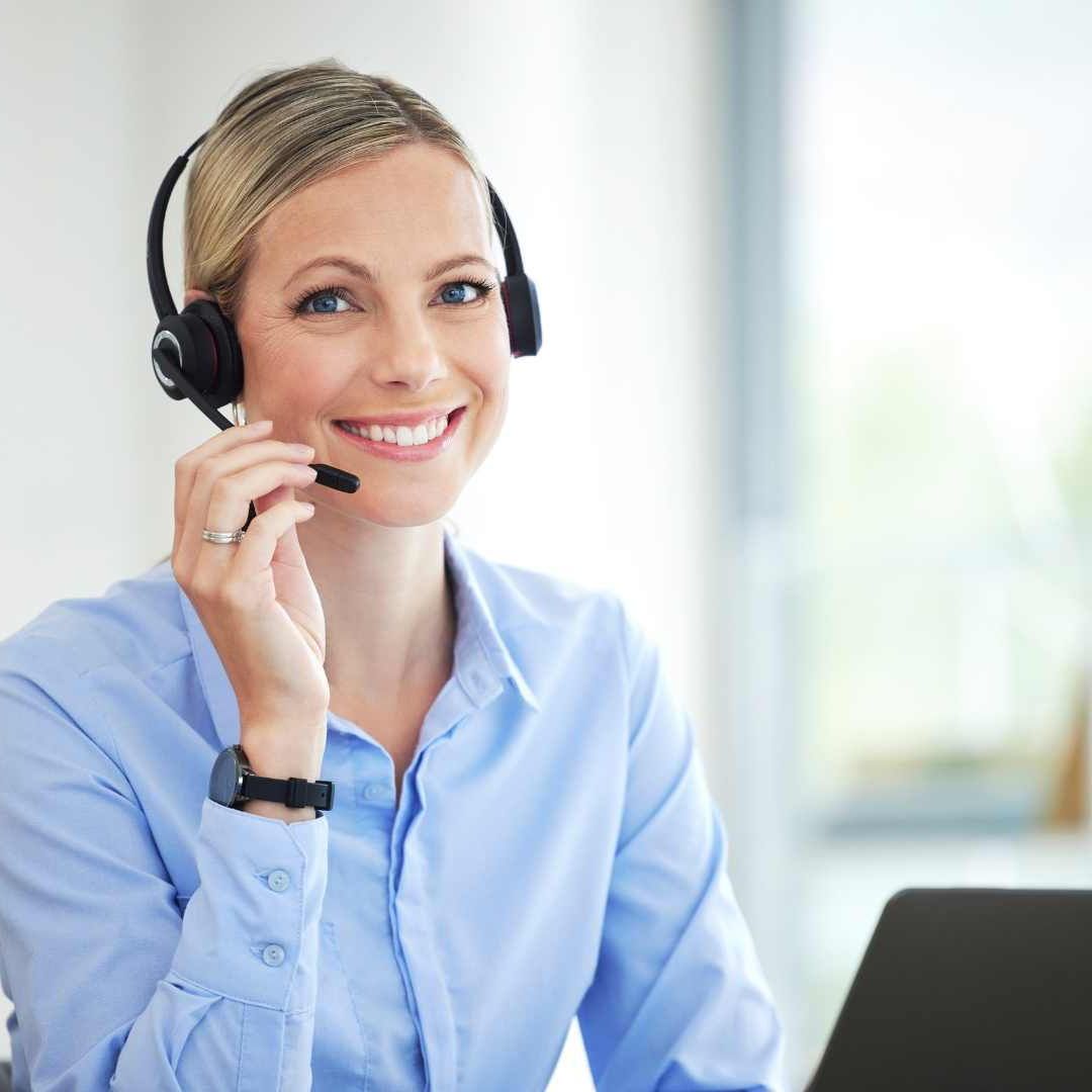 Blonde woman with headset smiles, holding microphone, wearing blue shirt, working in office.