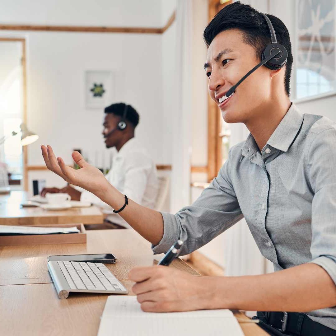 Man wearing headset gestures while speaking, writing notes in office, with another person in the background.