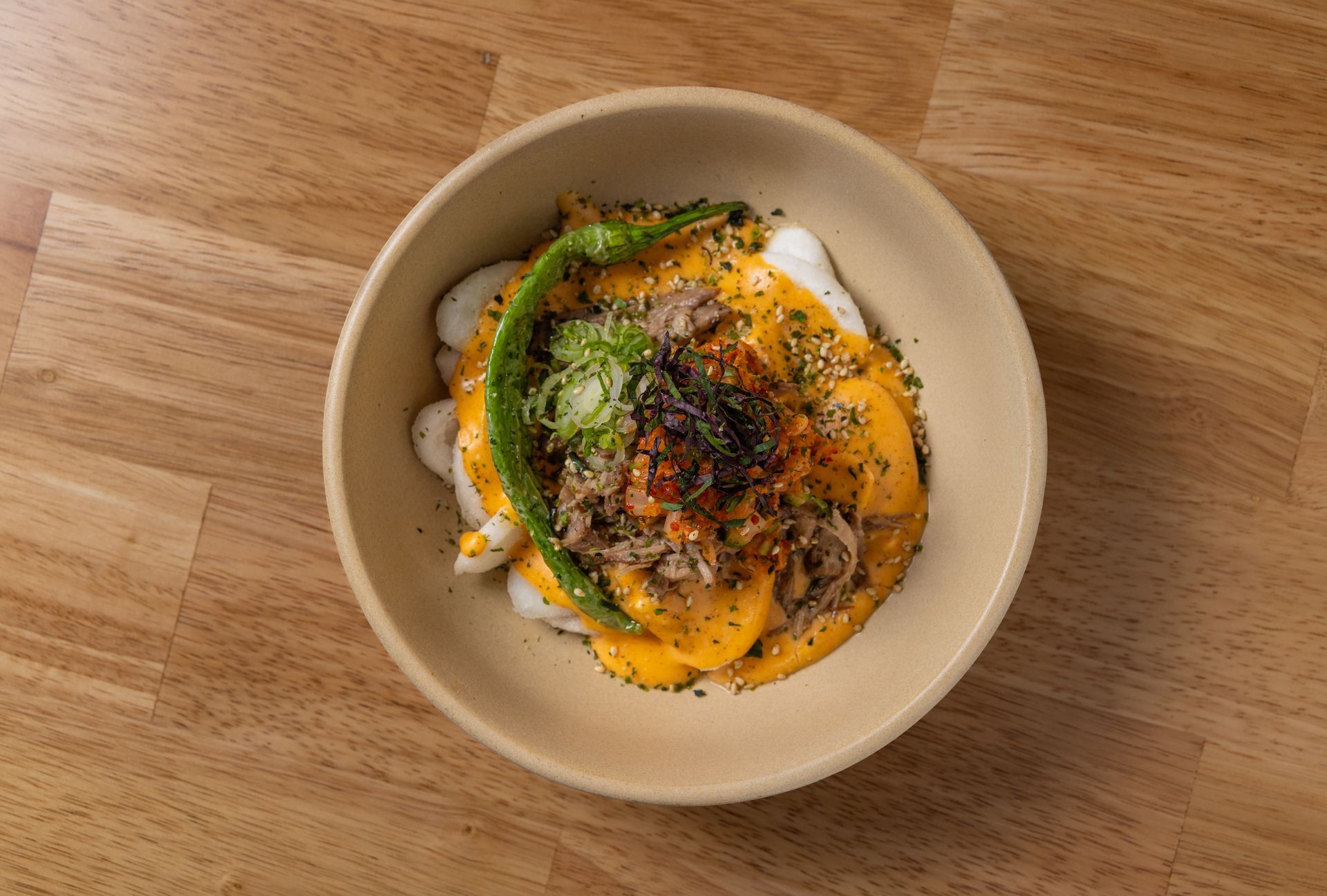 Bowl of food with rice cakes, sauce, meat, green pepper, and garnish on a wooden table.