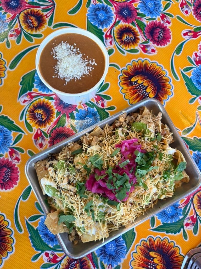 Tray of nachos and a bowl of beans on a floral tablecloth. Food is topped with cheese, cilantro, and pickled onions.