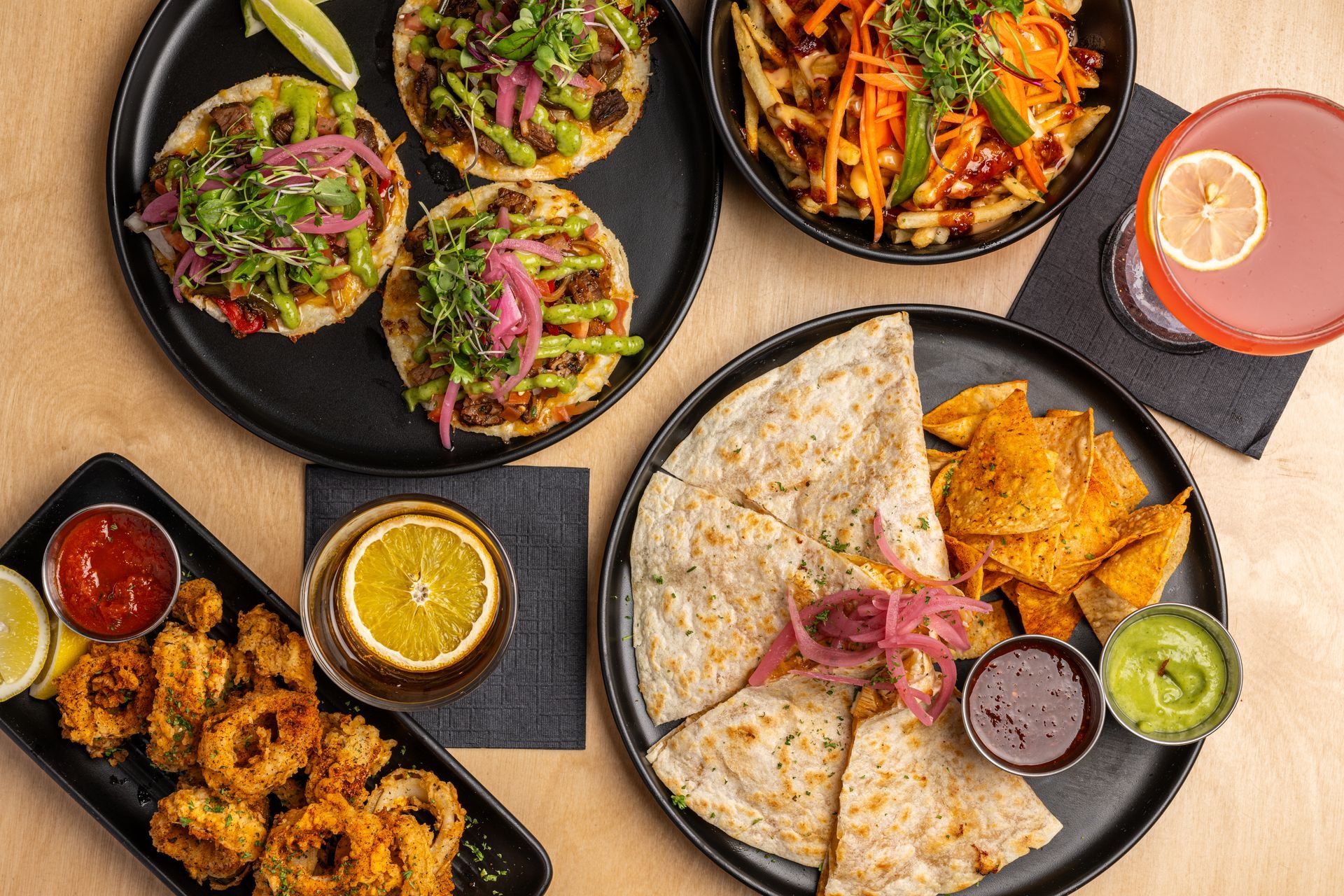 Overhead shot of a spread with tacos, fries, quesadilla, calamari, and cocktails on a wooden surface.