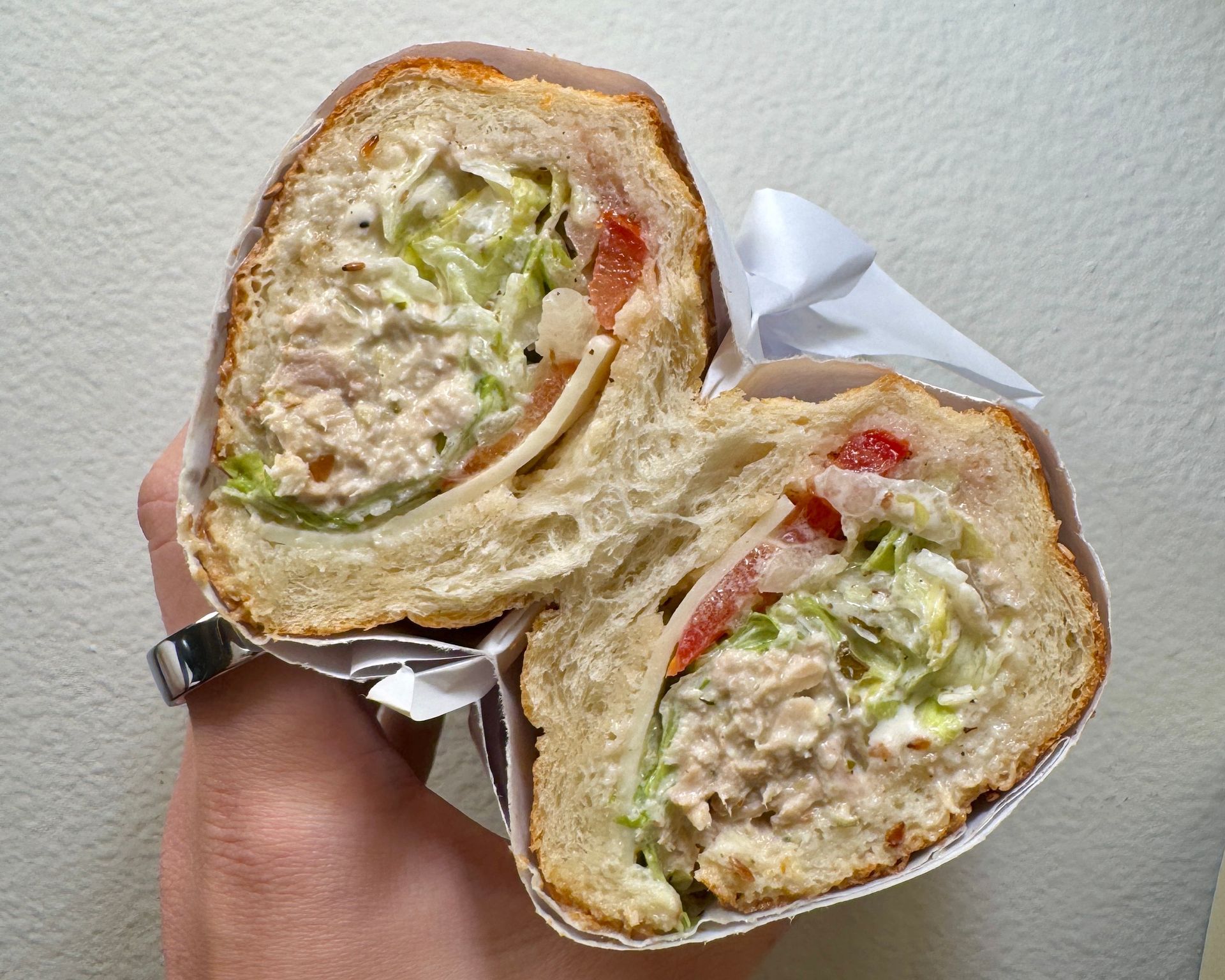 A halved tuna salad sandwich with lettuce, tomato, and bread. Held by a hand.