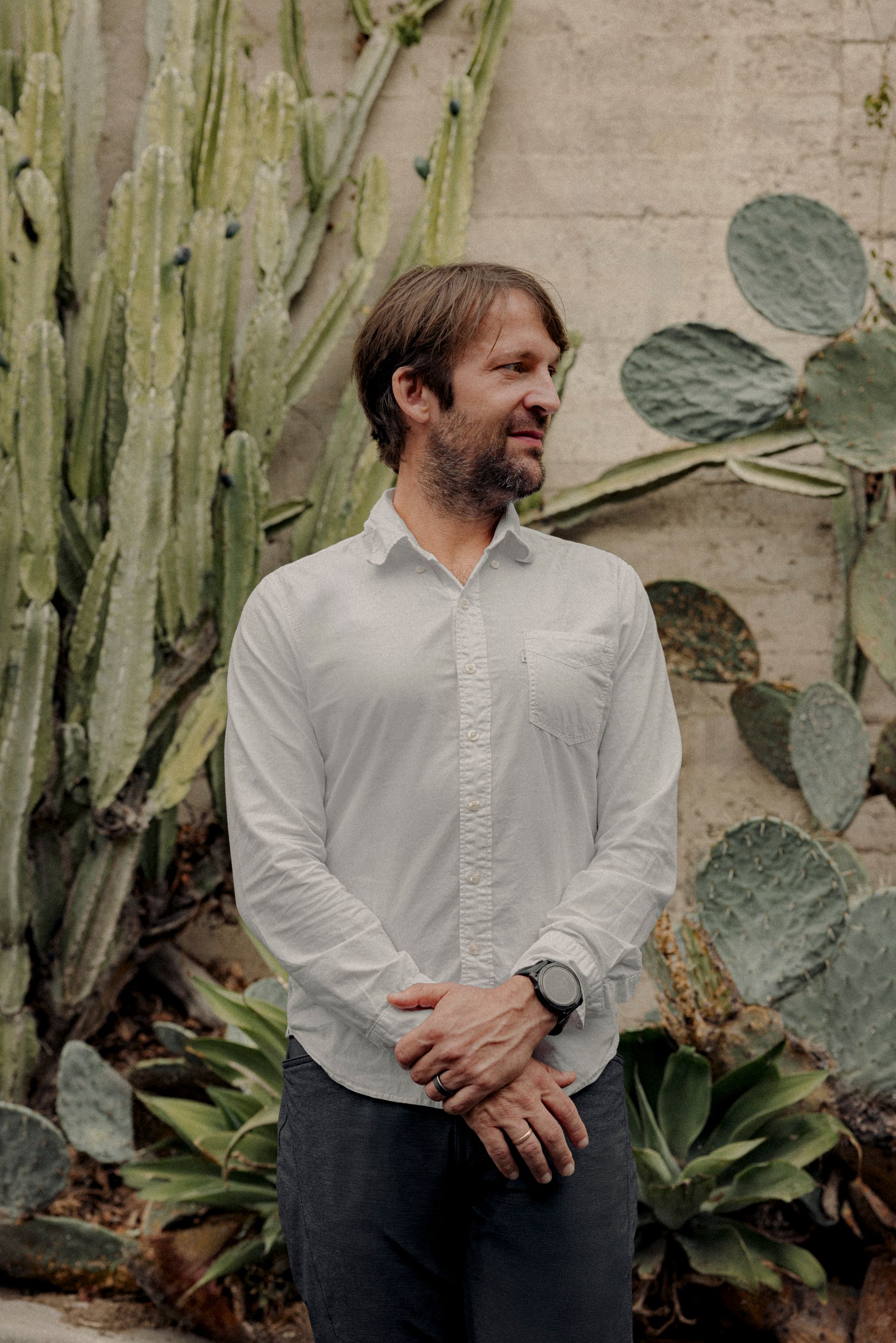 Man in white shirt and dark pants stands near cacti, looking to the side.