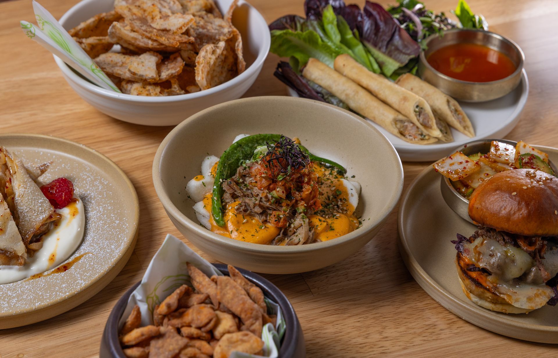 Various dishes of food, including spring rolls, a burger, and a bowl of salad, arranged on a wooden table.