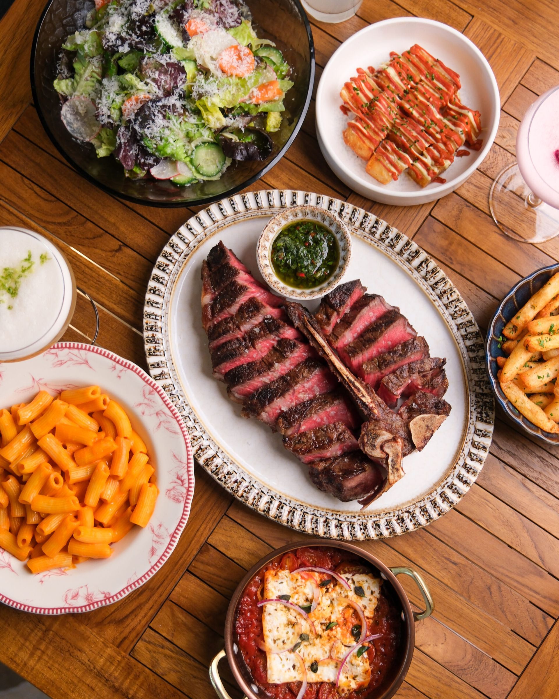 Overhead shot of a table set with steak, pasta, salad, fries, and cocktails.