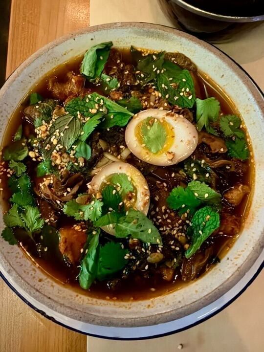 Bowl of dark broth with eggs, herbs, and sesame seeds.