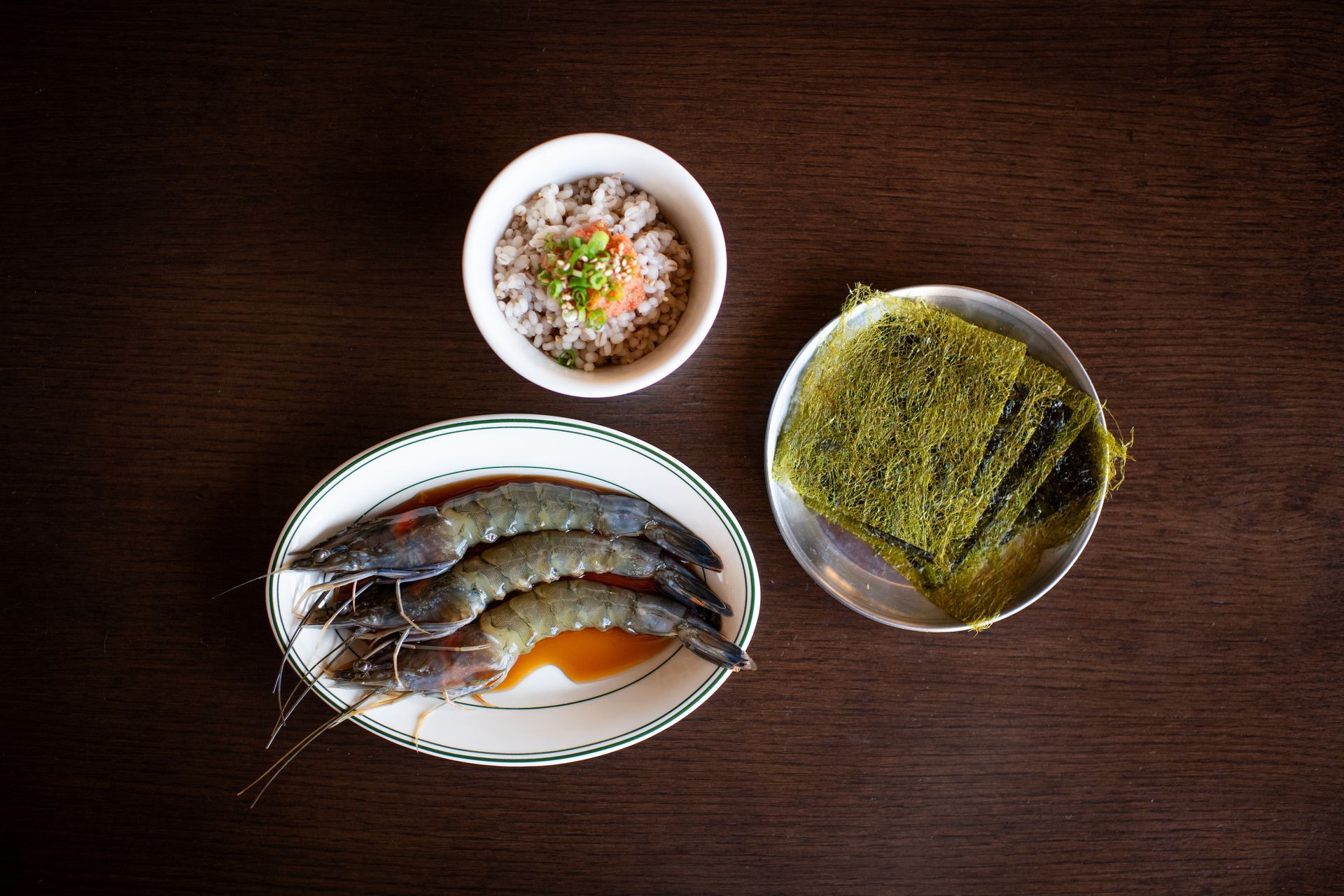 Plate of shrimp, bowl of topping, seaweed sheets on a dark wooden table.