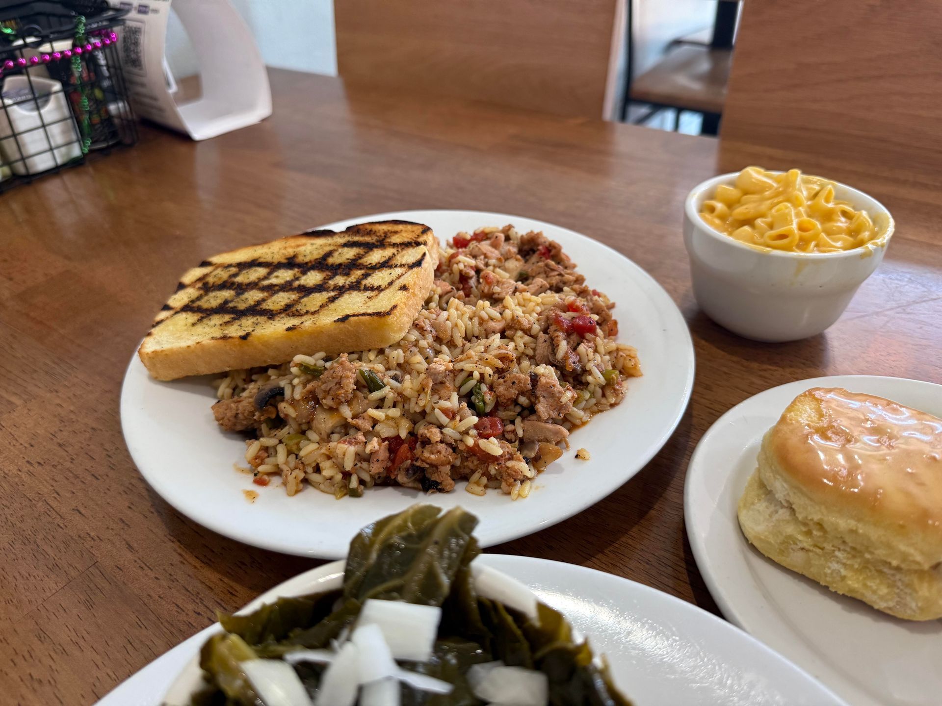 Plate with rice, sausage, and toast, with sides of mac and cheese, biscuit, and collard greens on a table.