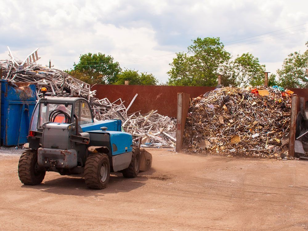 Blue forklift at a scrapyard with piles of metal and rusted debris near a fence.