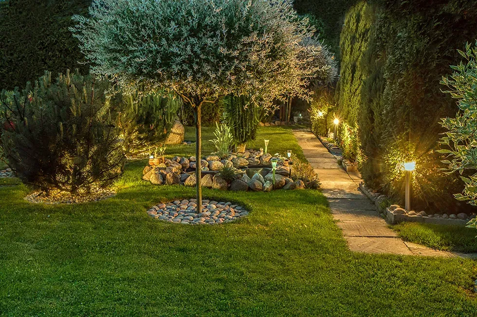 Illuminated garden path with lawn, flower beds, and trees at dusk