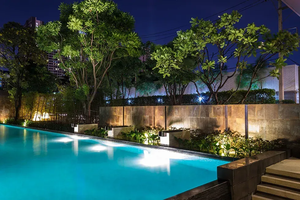 Illuminated resort pool at night with trees, stone wall, and steps along the edge
