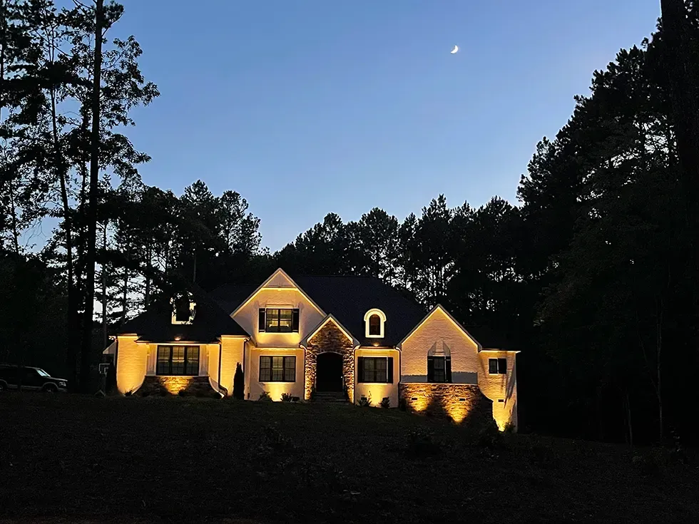 Illuminated house at dusk with warm lights, dark trees framing the scene, and a moon in the sky