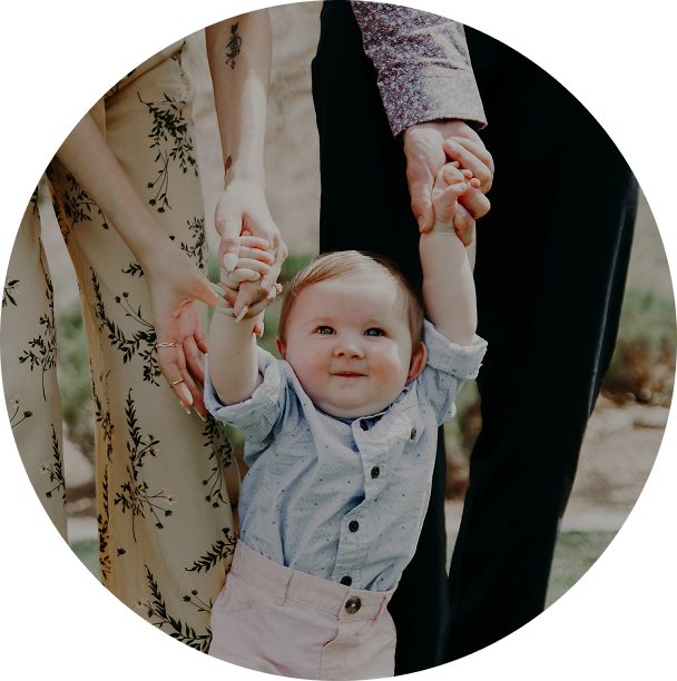 Toddler smiling while holding two adults’ hands outdoors, wearing a light blue shirt and beige pants.