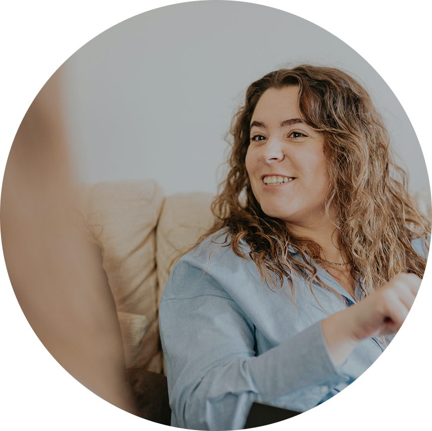 Smiling person with curly hair sitting on a beige couch in a light blue shirt