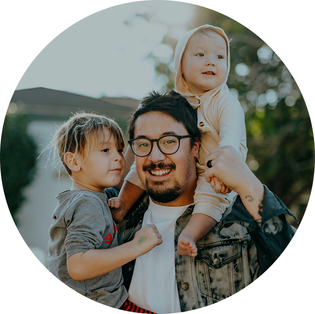 Adult smiling with two small children outdoors, one on shoulders, in a circular portrait