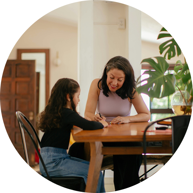 Woman and child sit at a wooden table, smiling and writing together in a bright home dining room