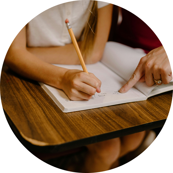 Hands writing in a notebook on a wooden desk, with one finger pointing to the page.