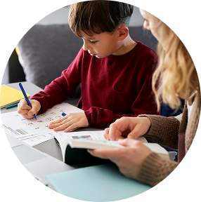 Child writing at a table while an adult reads beside them, with papers and a notebook in view.