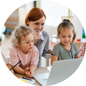 Teacher helping two children use a laptop at a table in a bright classroom