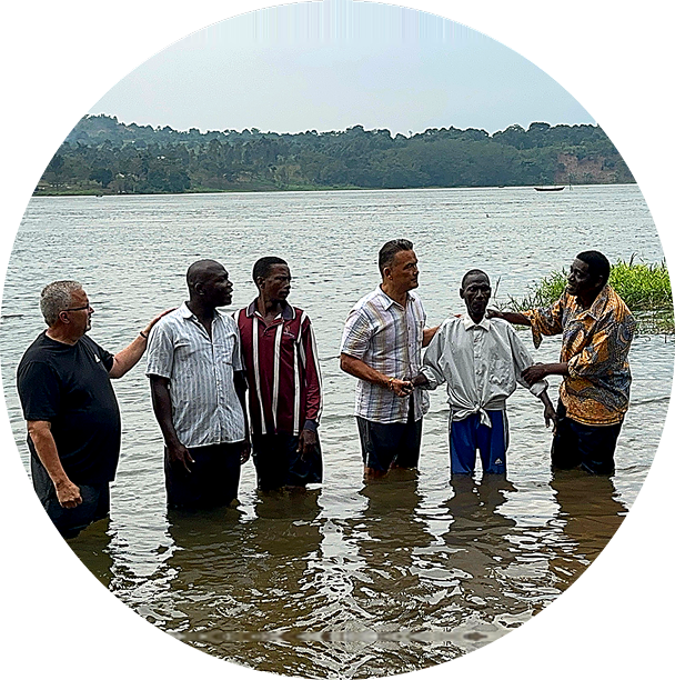 Six men standing in shallow water by a lakeside, talking near a wooded shoreline.